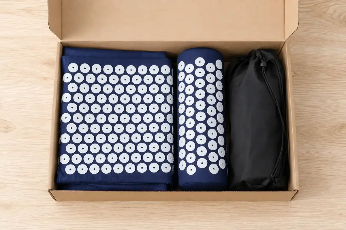 Open kraft box on a light wood table containing a folded navy SpikeRest mat, its bolster pillow and a black carry bag