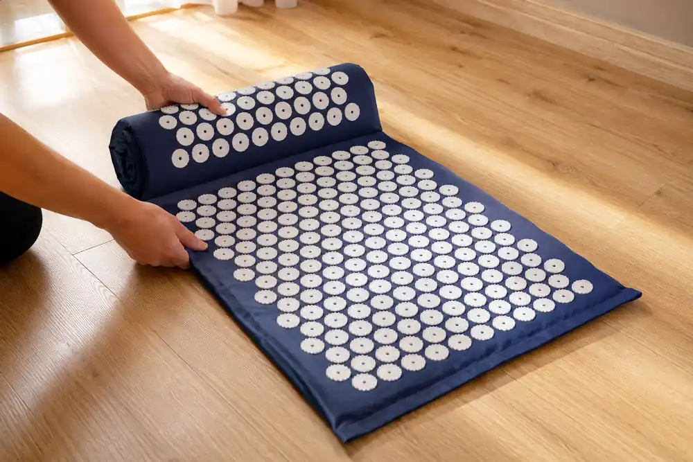 Two hands unrolling a navy acupressure mat onto a wooden floor