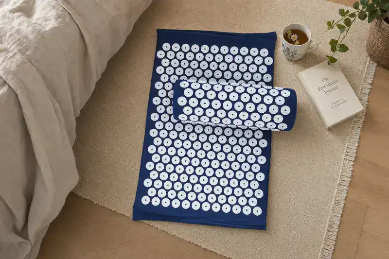 Navy blue acupressure mat with its matching bolster pillow laid flat on a rug beside a bed, next to a cup of tea and a book