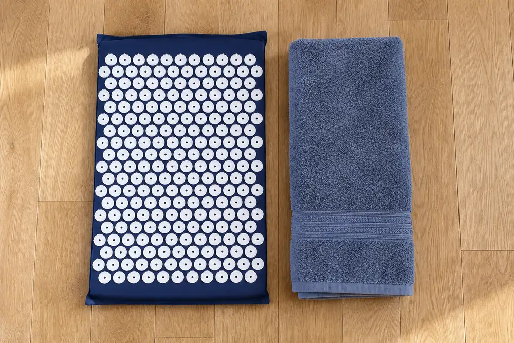 Navy SpikeRest acupressure mat laid on a wooden floor beside a folded bath towel of similar length, showing how compact it is